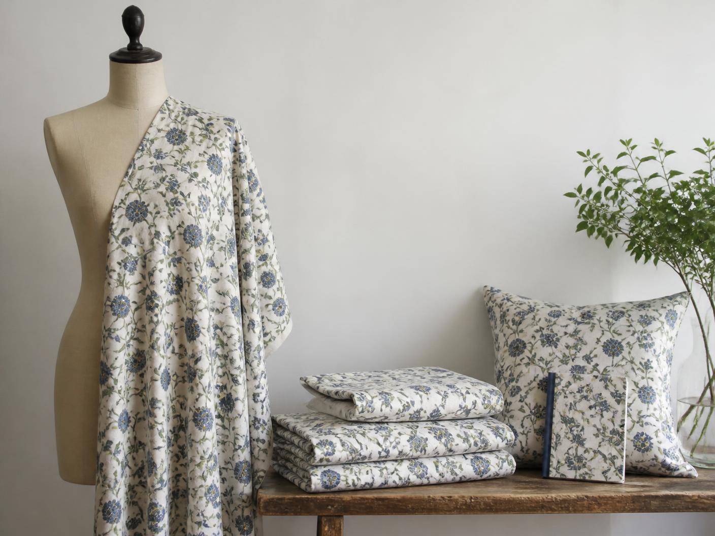 One botanical print shown across a draped dress length on a mannequin, folded bed linen, a cushion and a small notebook