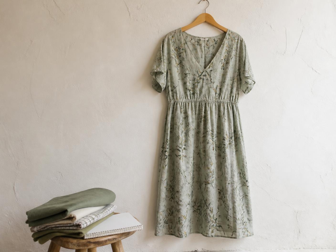 A sage botanical print organic cotton dress on a wooden hanger against a plaster wall, with folded fabric samples and a notebook on a stool