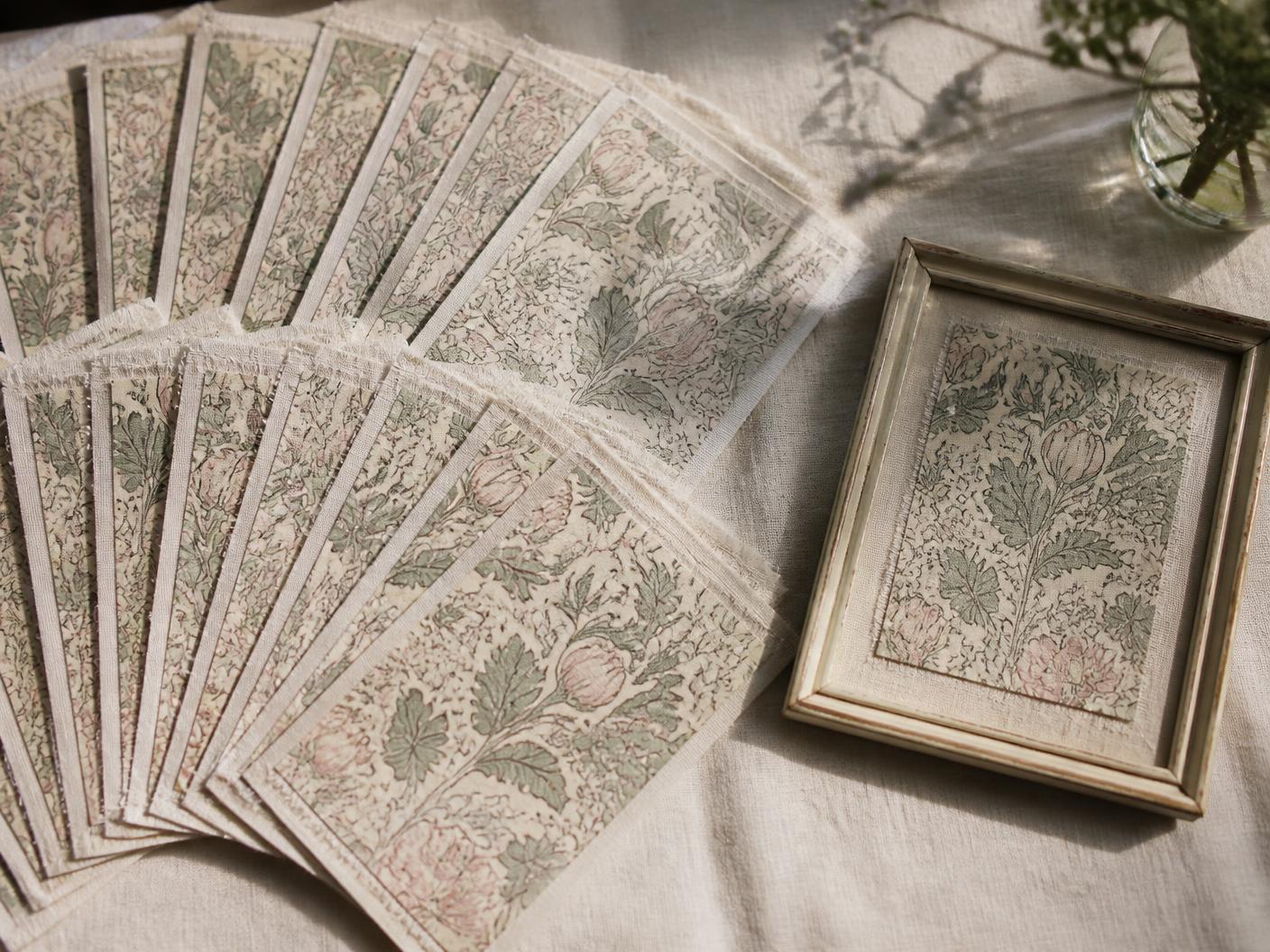 Many identical printed botanical fabric swatches fanned across a linen table beside one single framed swatch set apart