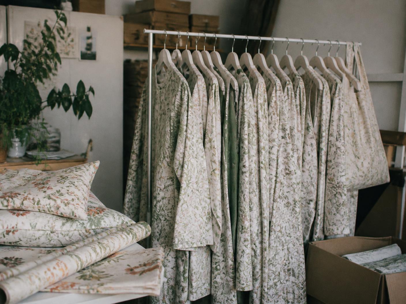 Small brand studio with a rail of printed botanical garments beside stacked printed cushions and rolls of patterned paper