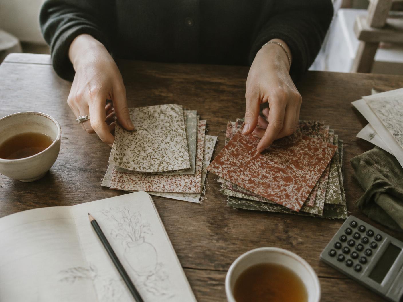 Designer's hands sorting printed fabric swatches into two piles on a wooden studio table beside a sketchbook and calculator
