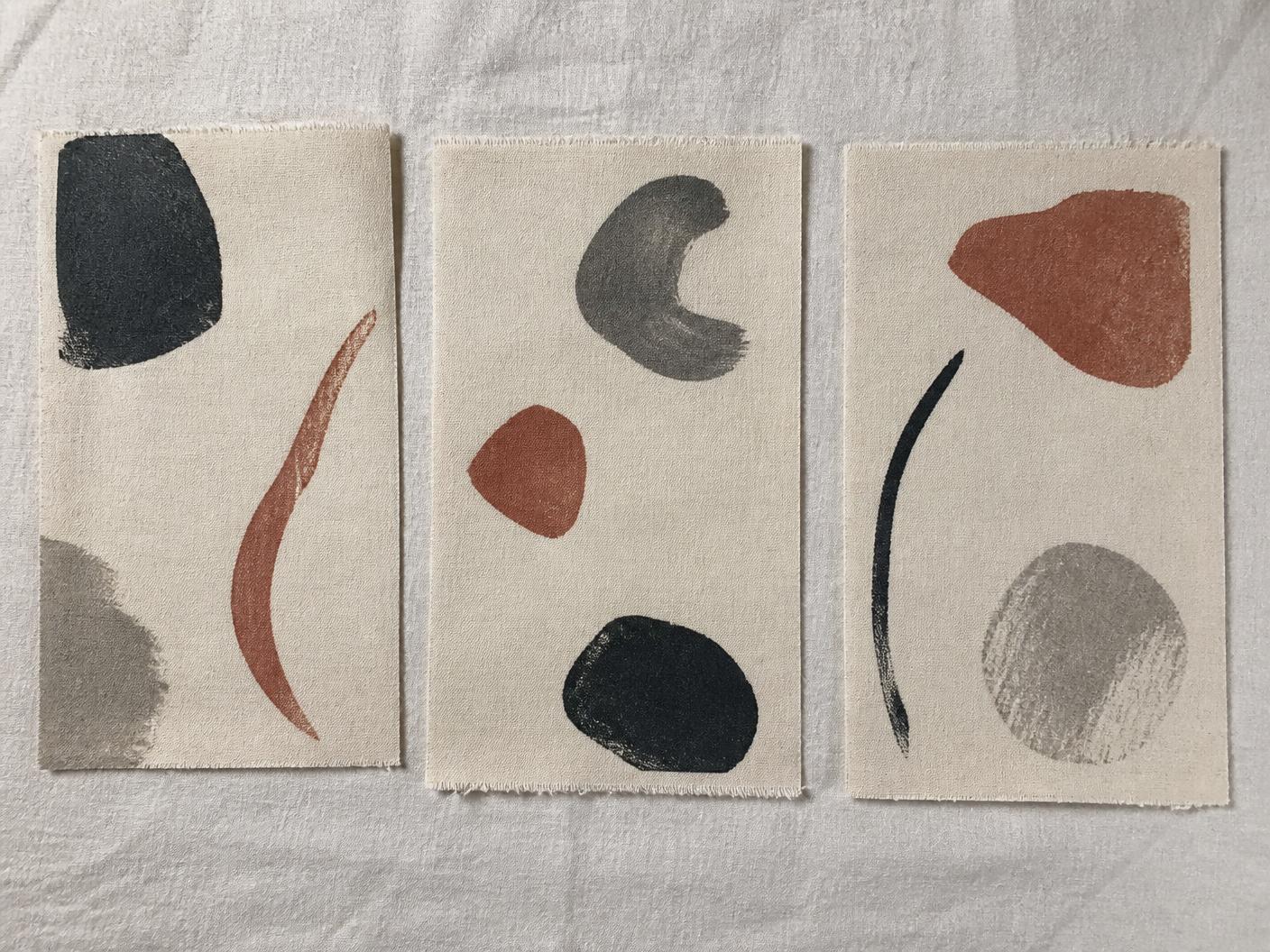 Three printed cotton swatches on pale linen, each with a few sparse abstract brush marks in charcoal, clay and grey on an ecru ground