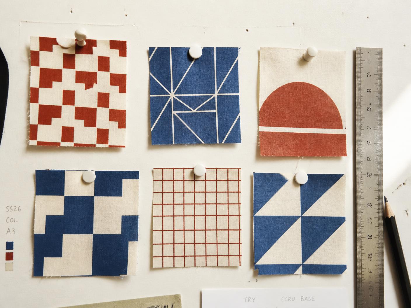 Six geometric printed cotton swatches in brick red, indigo blue and ecru pinned to a studio board beside a steel ruler and pencil