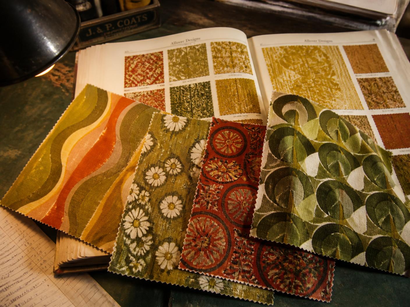 Vintage fabric swatches in avocado green, burnt orange and harvest gold layered over an open archive book of old textile samples under a desk lamp