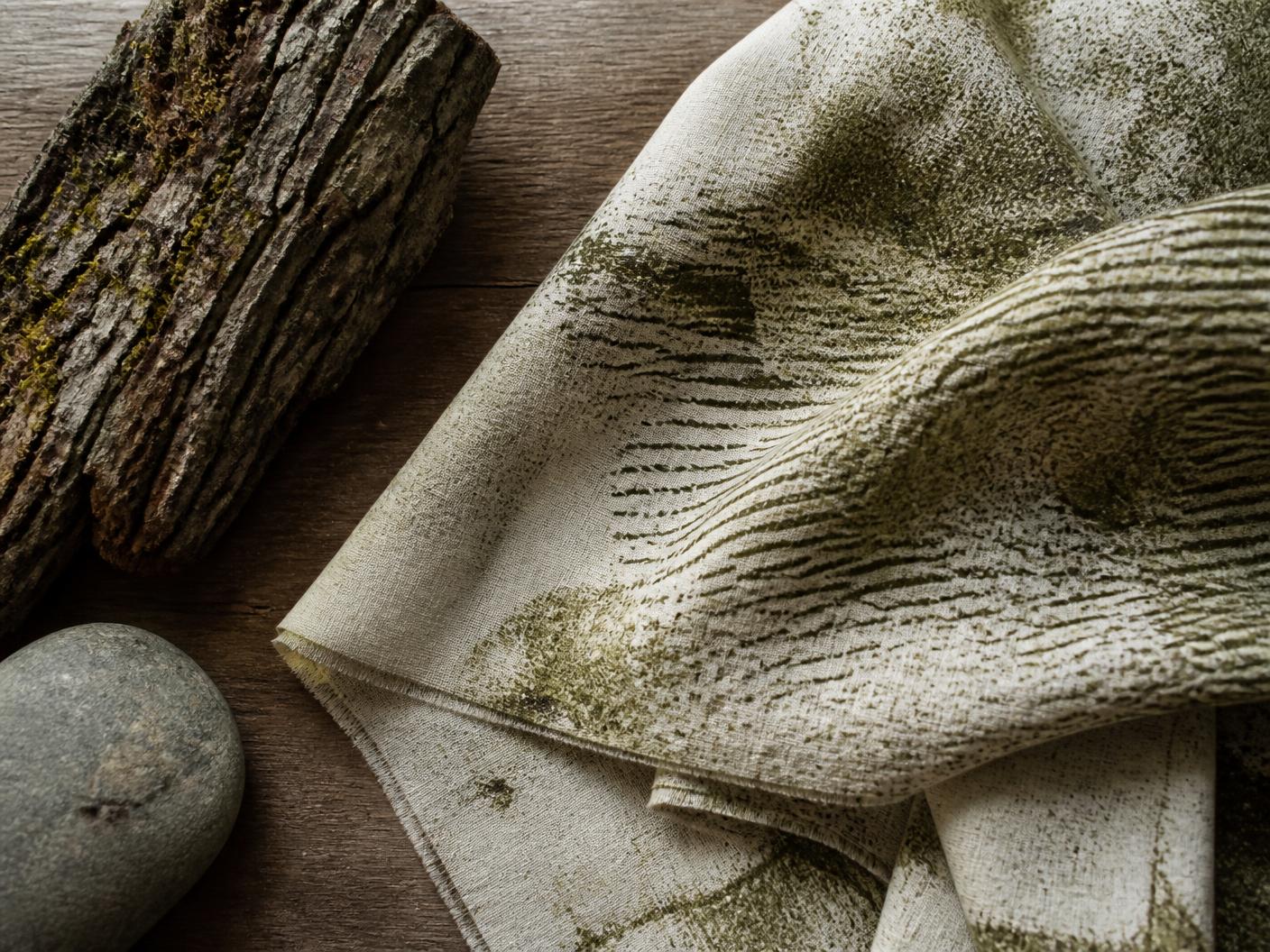Folded linen printed with abstract bark and stone textures in olive and cream, laid on a wooden table beside a piece of bark and a grey stone